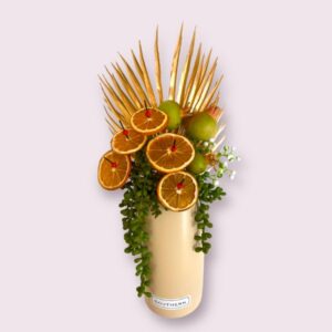 A long life abstract arrangement with gold fan leaf, pommegranite, hanging green foilage, preserved oranges in a tall sand coloured vase
