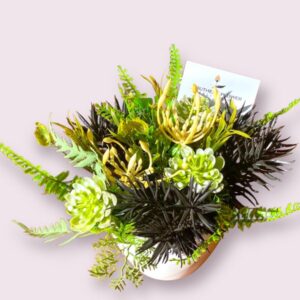 An under the sea themed long life arrangement with varying shades of green to represent underwater plants with a contrast of black sea urchins in a white boat shaped vase