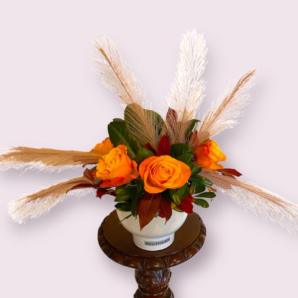 Orange roses, splashes of red amongst leaves and mauve reeds in an elegant smoke grey coloured bowl vase.