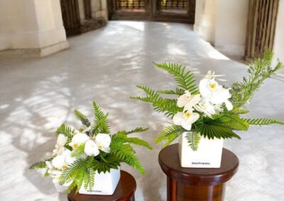 Two small white vases with white orchids and greenery pictured with two art deco brown timber pedestals all available for hire