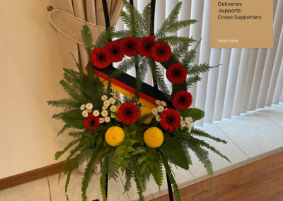 Picture of wreath with red gerberas, ribbons of the crows colours, yellow contrast flowers amongst green foilage on a black stand